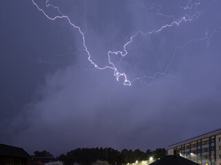 A dramatic night sky lit up by vivid lightning bolts during a thunderstorm, with buildings and a...