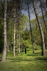 Obraz premium A lone girl in a dark dress stands in a sun-dappled clearing surrounded by tall pine trees, casting long shadows across the grassy forest floor on a bright day