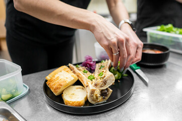 A chef's hand garnishes a plate with roasted bone marrow, fresh herbs, and crispy bread. The meticulous arrangement showcases culinary artistry in a professional kitchen setting.