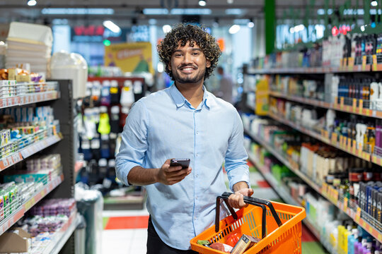 A smiling man with a shopping basket and a phone browses the aisles of a brightly lit grocery store, preparing to buy groceries.