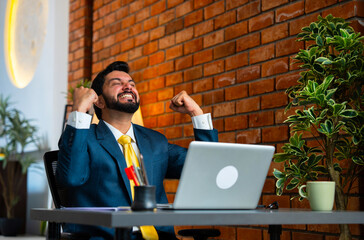 Smiling Indian businessman celebrates a big win while working on laptop in cozy office setup