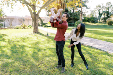 Smiling mom standing behind dad holding little girl upside down