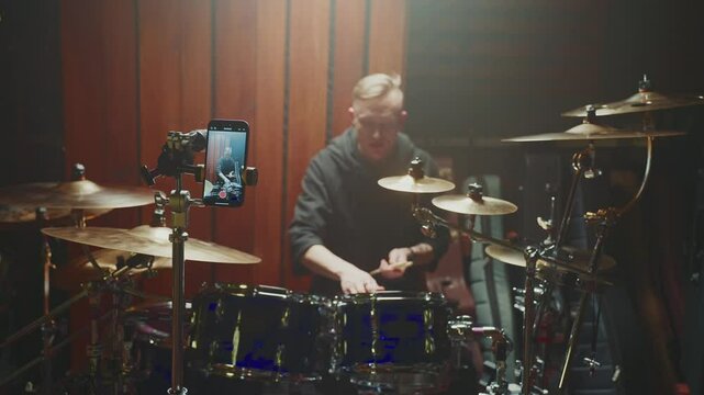 A musician plays an energetic drum solo in a studio, showcasing skill and passion. A smartphone records the performance, capturing the creative atmosphere and intense focus.