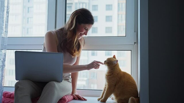 A woman enjoys quality time with her orange cat by petting it as she sits beside a window, working on her laptop in a bright and cozy apartment during the day.