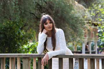 Naklejka premium Young and beautiful brunette woman in white knitted dress, makes different expressions leaning on the wooden railing of a bridge in the park. In the background trees and vegetation.