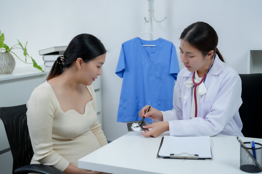 Asian pregnant woman consulting with female doctor during prenatal checkup receiving ultrasound explanation sitting together in medical office expressing health concern support and motherhood bonding