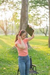 Asian woman sitting listening to music, reading a book, outdoors in nature, evening.