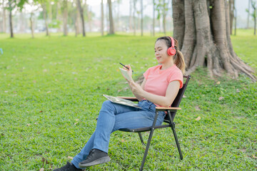 Asian woman sitting listening to music, reading a book, outdoors in nature, evening.