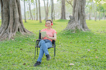 Asian woman sitting listening to music, reading a book, outdoors in nature, evening.