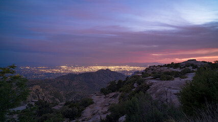 Vibrant colors paint the sky as night descends over a rocky terrain, revealing a distant Tucson shimmering with lights against the backdrop of a serene natural setting.