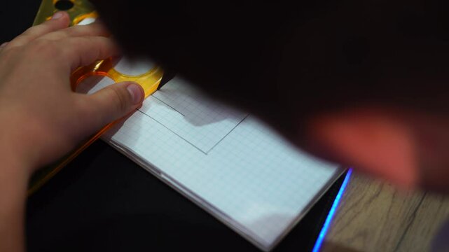 boy draws simple drawing of house with pencil and ruler in squared notebook. hand with pencil and triangle close-up