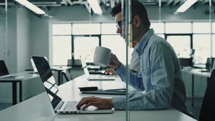 Professional developer focusing on project deadline while drinking coffee, working efficiently at organized desk with laptop in bright, modern office environment