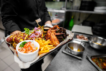 A chef holds a large platter filled with sausages, crispy chicken wings, golden fries, and fresh salad in a bustling kitchen. The vibrant presentation highlights the culinary artistry involved.