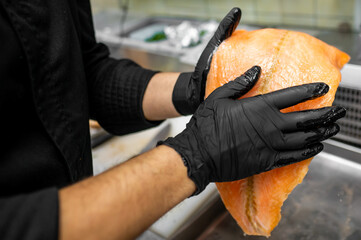 A chef in black gloves carefully handles a large piece of salmon, preparing it for cooking. The...