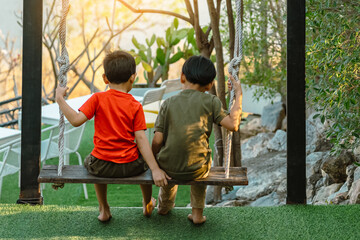 Back view of two Asian boys enjoy on swing. Brothers playing on wooden swing in park. Two little...