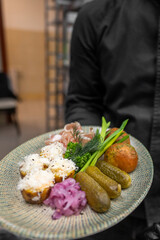 A chef presents a beautifully arranged plate featuring roasted potatoes, pickles, fresh herbs, and slices of ham. The artistic plating showcases vibrant colors and textures, enhancing visual appeal.
