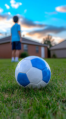 Obraz premium Blue and White Soccer Ball on Green Grass with Boy in Background at Sunset