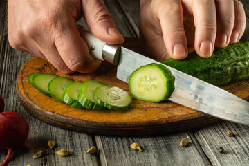Hands skillfully slice a fresh cucumber on a wooden cutting board. Bright green slices are placed...