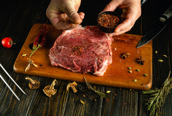 Hands expertly sprinkle aromatic spices over a thick cut of meat resting on a wooden board,...