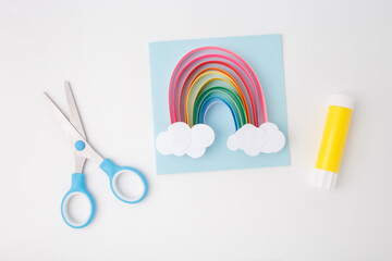 Colorful handmade paper rainbow with clouds on blue square, glue stick and scissors on white...