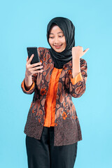 Smiling young Asian Muslim woman in batik outer and hijab pointing her thumb while holding a...