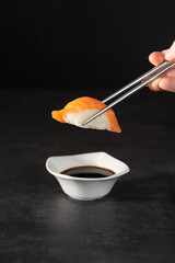 A hand holding a piece of salmon nigiri over a small bowl of soy sauce with stainless steel chopsticks. Black bacground.
