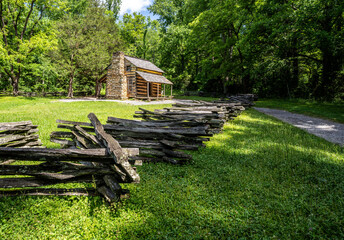 A log cabin with windows, a covered porch and a stone chimney on the edge of a forest with gravel road and a split rail fence, John Oliver Place, Great Smoky Mountain National Park, Tennessee