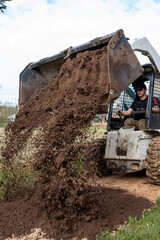 A worker uses a skid steer loader to dump loose soil onto the ground. The scene includes a wire...