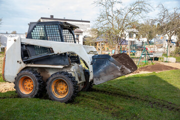 Obraz premium A skid steer loader with orange wheels carries soil in its raised bucket on a grassy surface. Nearby are houses, a swing set, and leafless trees.