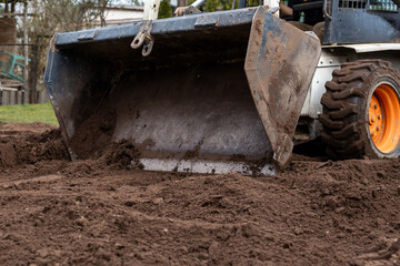 Close up of a white construction vehicle with orange wheels and a metal bucket moving soil. Dirt...