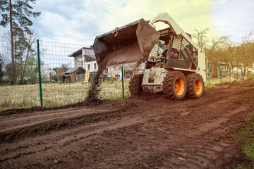 Fototapeta premium A skid steer loader with orange wheels spreads soil on a dirt path near a wire fence, with residential buildings and sparse trees in the distance.