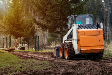 An orange and white skid steer loader parked on a dirt path with visible tire tracks, bordered by a metal fence and surrounded by tall evergreen trees. © True Pixel Art