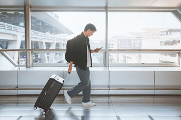 Side view - Male traveler walking with luggage using mobile smartphone at the modern airport terminal, man on way to flight boarding gate, Ready for travel or vacation journey, copy space