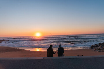 Two fishermen fishing on the ocean shore with sunset in the background