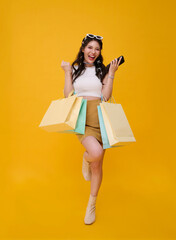 Happy celebrating cute Asian woman shopper carrying colorful shopping bags isolated on yellow...