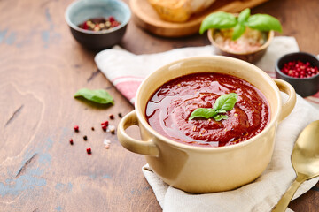 Tomato puree soup garnished with basil leaves and spices in ceramic bowl with toasted bread slices on wooden table