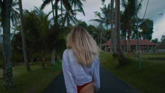 A sexy girl model in the tropics is walking along the road among palm trees. Beautiful woman in Asia is flirting in a white shirt. Blonde female enjoys a vacation in bali indonesia. Perfect ass