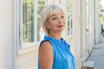 A stylish woman aged 65-70 in a blue outfit stands outside a building with window