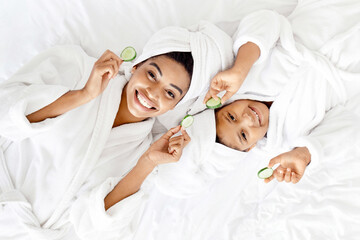 Home Spa. Top View Of Happy African Mom And Daughter In Bathrobes And Towels With Cucumber Slices Doing Face Mask After Bath, Having Beauty Day, Lying On Bed, Smiling At Camera, Copy Space