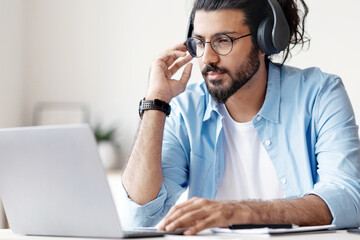 Distance Learning. Young Eastern Man In Headphones Studying Online With Laptop At Home, Watching Webinar Or Training Lesson In Internet, Sitting At Desk And Looking At Computer Screen, Free Space