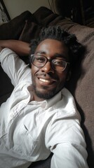 Fototapeta premium A cheerful man lounges comfortably on a soft couch, wearing a white shirt, emanating warmth and relaxation. Sunlight gently illuminates the cozy space, enhancing his joyful expression