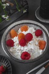 An easy healthy breakfast. A bowl filled with grainy, creamy cottage cheese is decorated with bright yellow and red raspberries. The dish stands on a dark textured surface. Close-up
