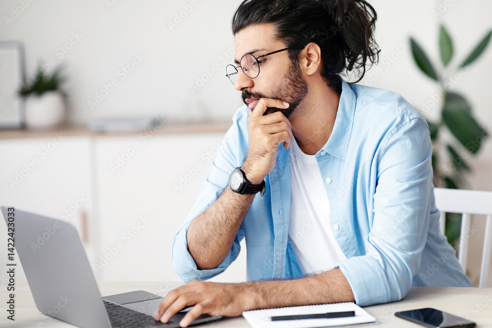 Poster thoughtful indian freelancer working with laptop at home office, sitting at desk looking at computer