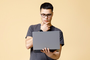 Designer and freelancer works online. Pensive serious young man in brown t-shirt rubs his chin and...