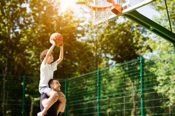 Healthy Lifestyle. Curly son sitting on his daddy's shoulders, throwing ball into basket on sunny...