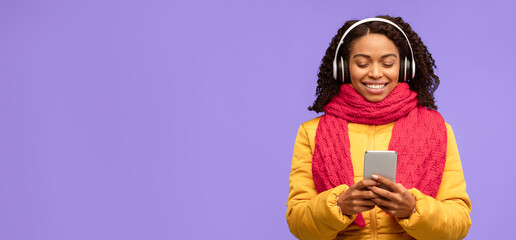 Young Black Woman Listening To Music On Phone Enjoying Favorite Song Standing Over Blue Studio Background, Wearing Warm Jacket. Musical Mobile App And Winter Playlist Concept. Panorama, Free Space