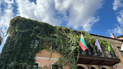 Charming Hotel Facade In Plovdiv, Bulgaria, Covered In Lush Green Vines With Multiple Flags Including The Bulgarian National Flag On A Balcony. Clear Blue Sky And Clouds In The Background.