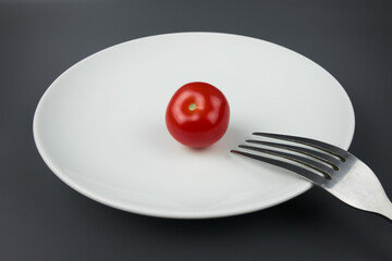 A small red cherry tomato on a white plate with a fork, gray background, close-up. The concept of dietary healthy eating, weight loss and vegetarianism.