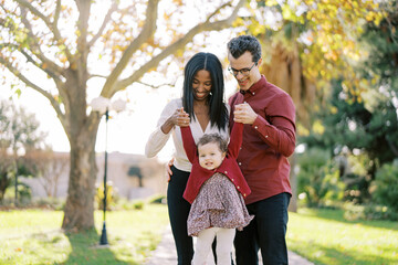 Fototapeta premium Smiling parents raising little girl by hands while standing in park