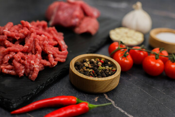 Close-up on a dark background in focus of peppercorns in a wooden bowl and shredded red minced meat with streaks of fat and red hot pepper surrounded by vegetables, meat and seasonings